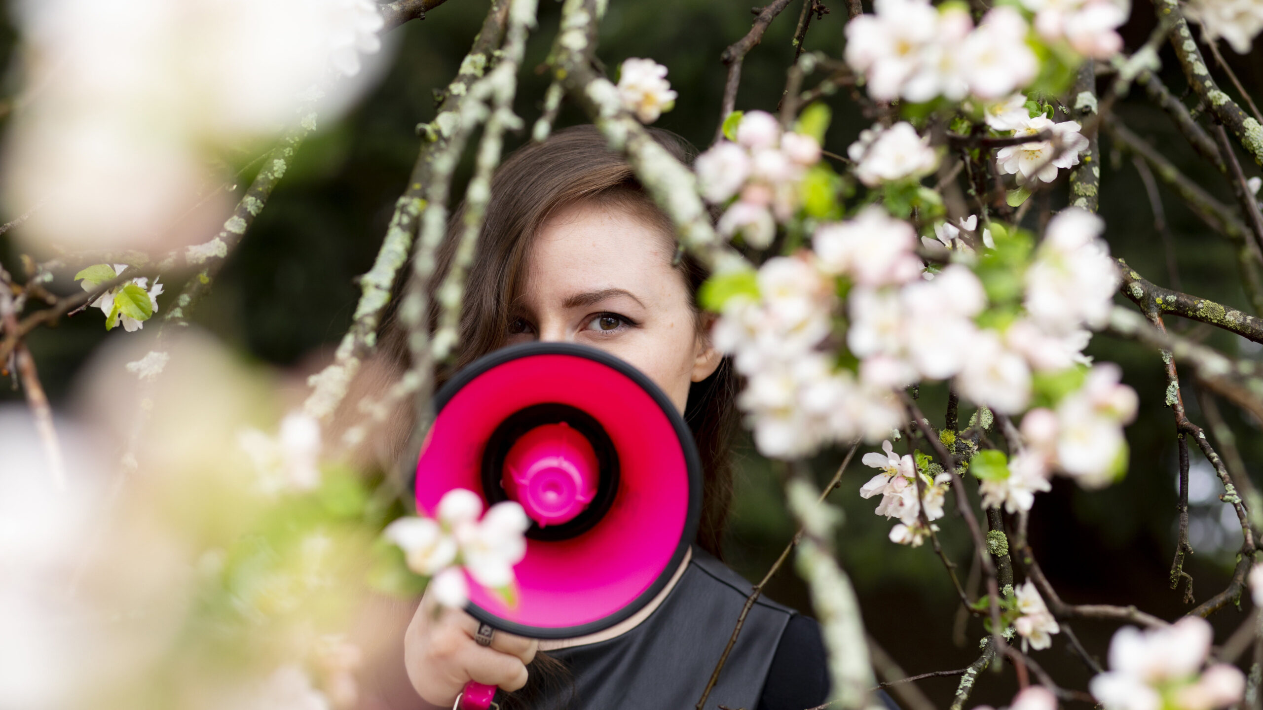 cover image showing Tina behind a pink megaphone looking directly at the camera, with her face partially obscured by the megaphone so that only her eye is visible. she is surrounded by branches of a flowering tree with a narrow plane of focus so that the branches in the foreground are blurry.